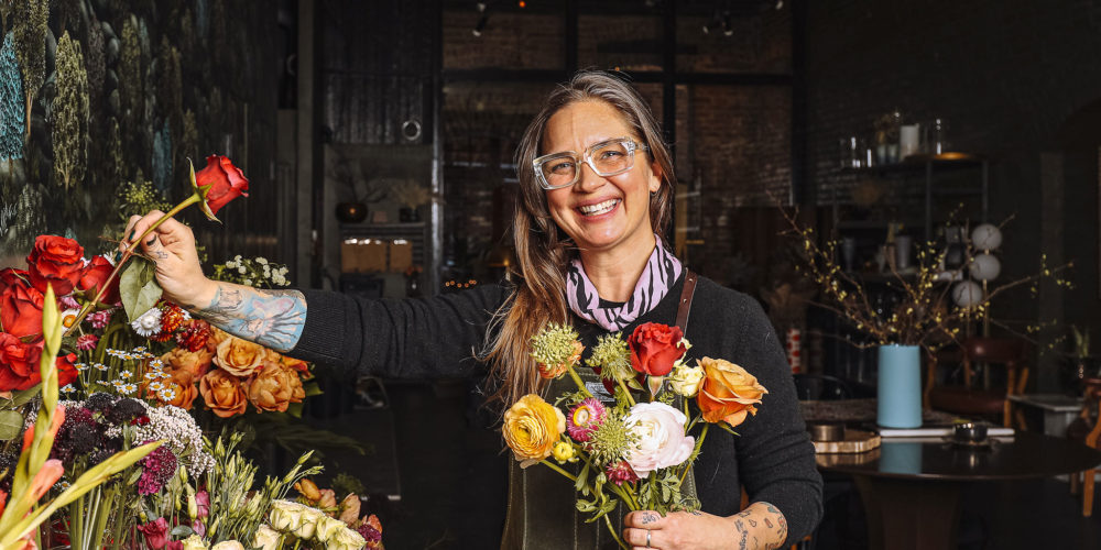 Becky stands in her floral shop holding a bouquet of flowers, smiling.