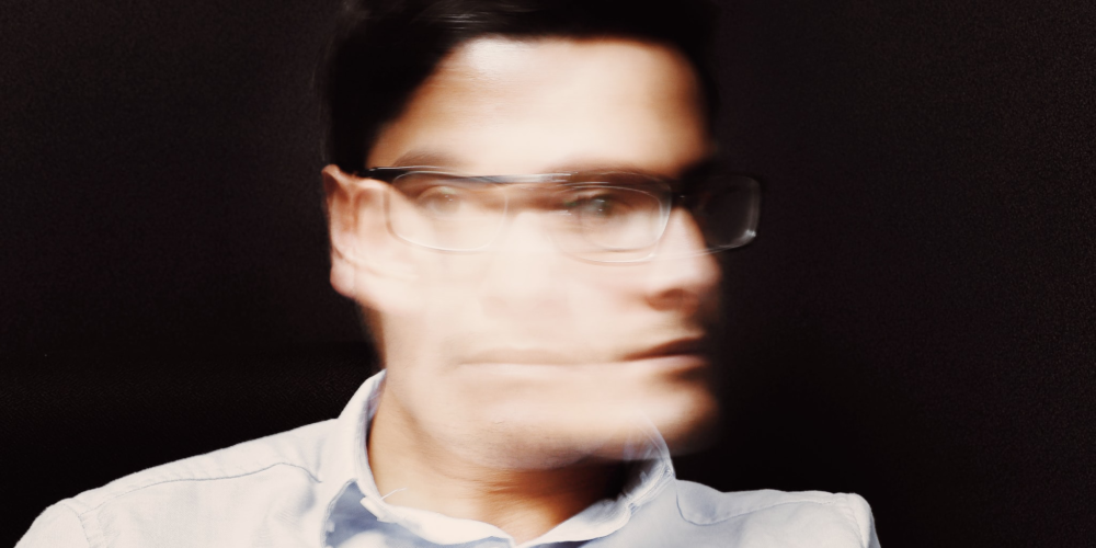 A blurry image of a man wearing glasses and shaking his head.