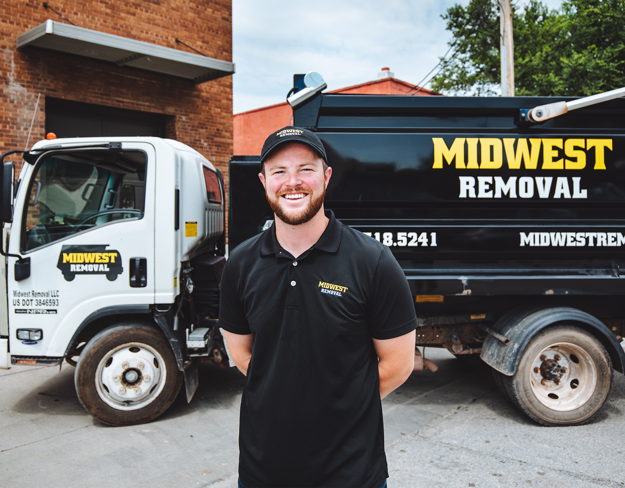 A man, Joe Manzari, wearing a black polo and pants stands smiling in front of a Midwest Removal tuck.