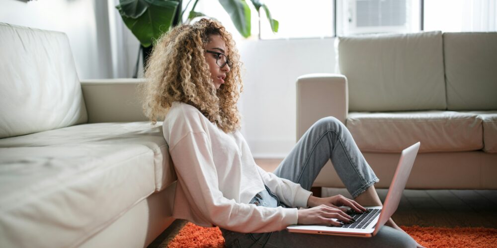 A woman sitting on a orange rug leaning against a white couch. She has curly hair and is wearing a white long sleeved shirt and jeans. She is working on a laptop computer that is resting on her leg.