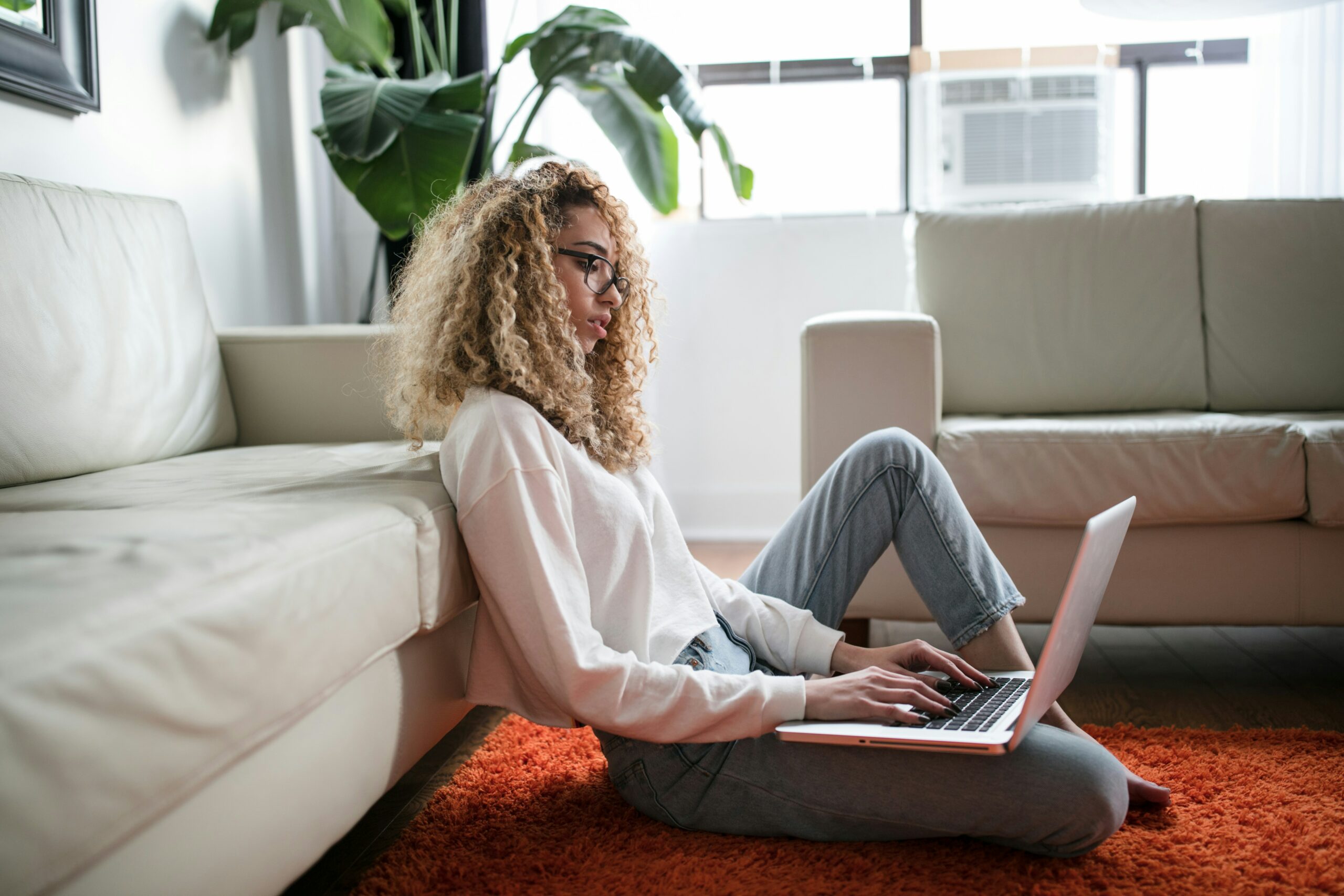 A woman sitting on a orange rug leaning against a white couch. She has curly hair and is wearing a white long sleeved shirt and jeans. She is working on a laptop computer that is resting on her leg.