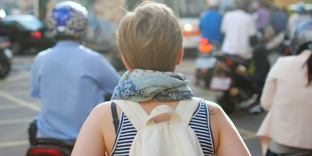 From behind: We see a woman with short dirty blonde hair walking through a crowded street. She is wearing a white backpack and there is a person on a motorcycle just in front of her.