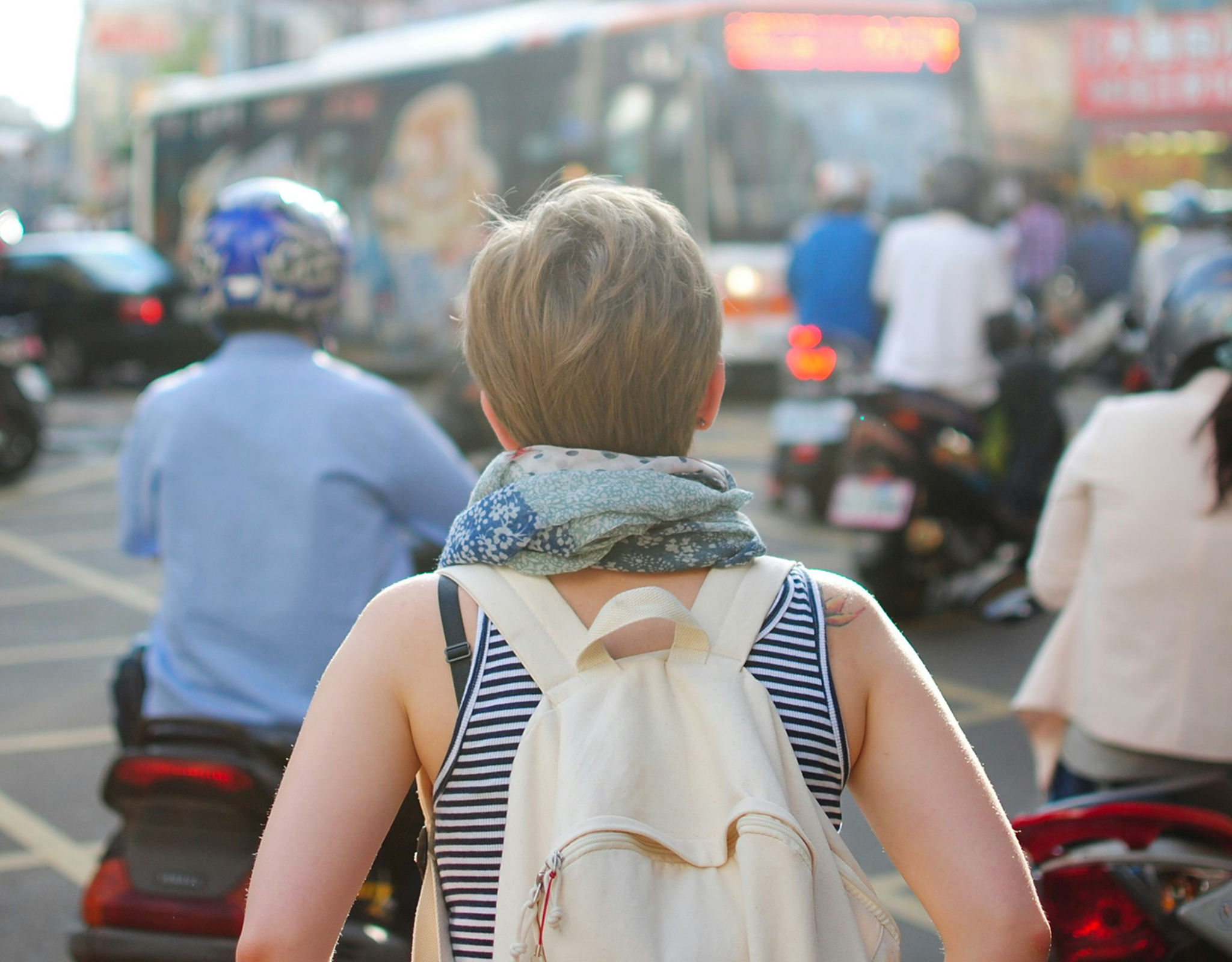From behind: We see a woman with short dirty blonde hair walking through a crowded street. She is wearing a white backpack and there is a person on a motorcycle just in front of her.