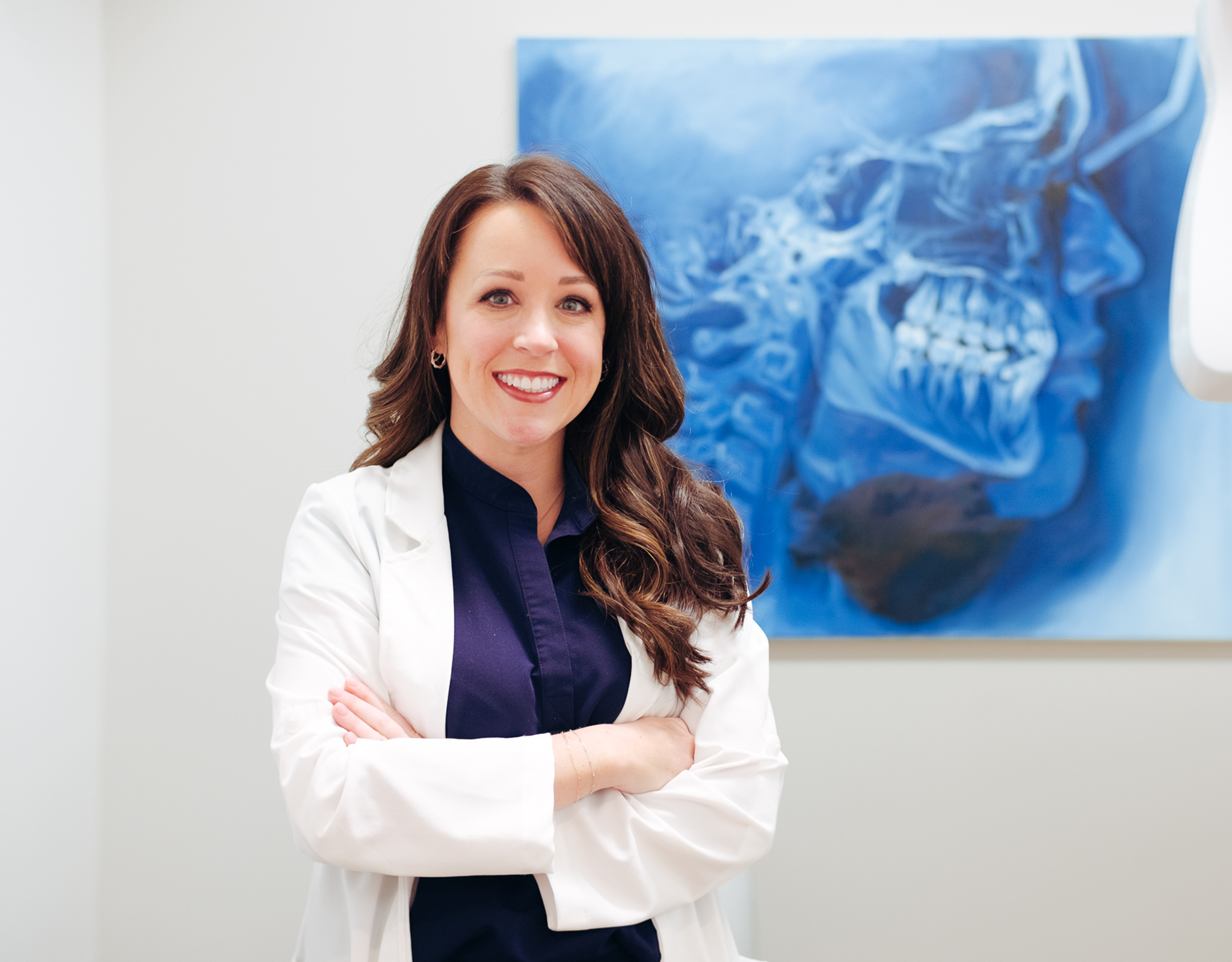 A woman, Dr. Christina Pruitt, stands smiling at the camera. An large x-ray of human jaw/teeth hangs on the wall behind her.